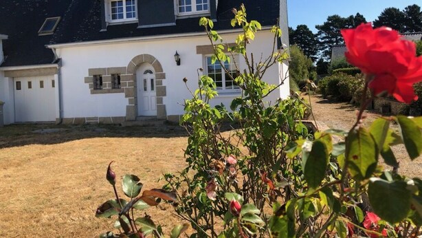 Una casa de dos pisos con fachada blanca, techo oscuro y un pequeño jardín con una rosa roja en primer plano.