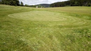 A large, open grassy field with a circular pattern, surrounded by trees and hills in the background. - Garden & Pets Escape by Interhome (Tanvald)