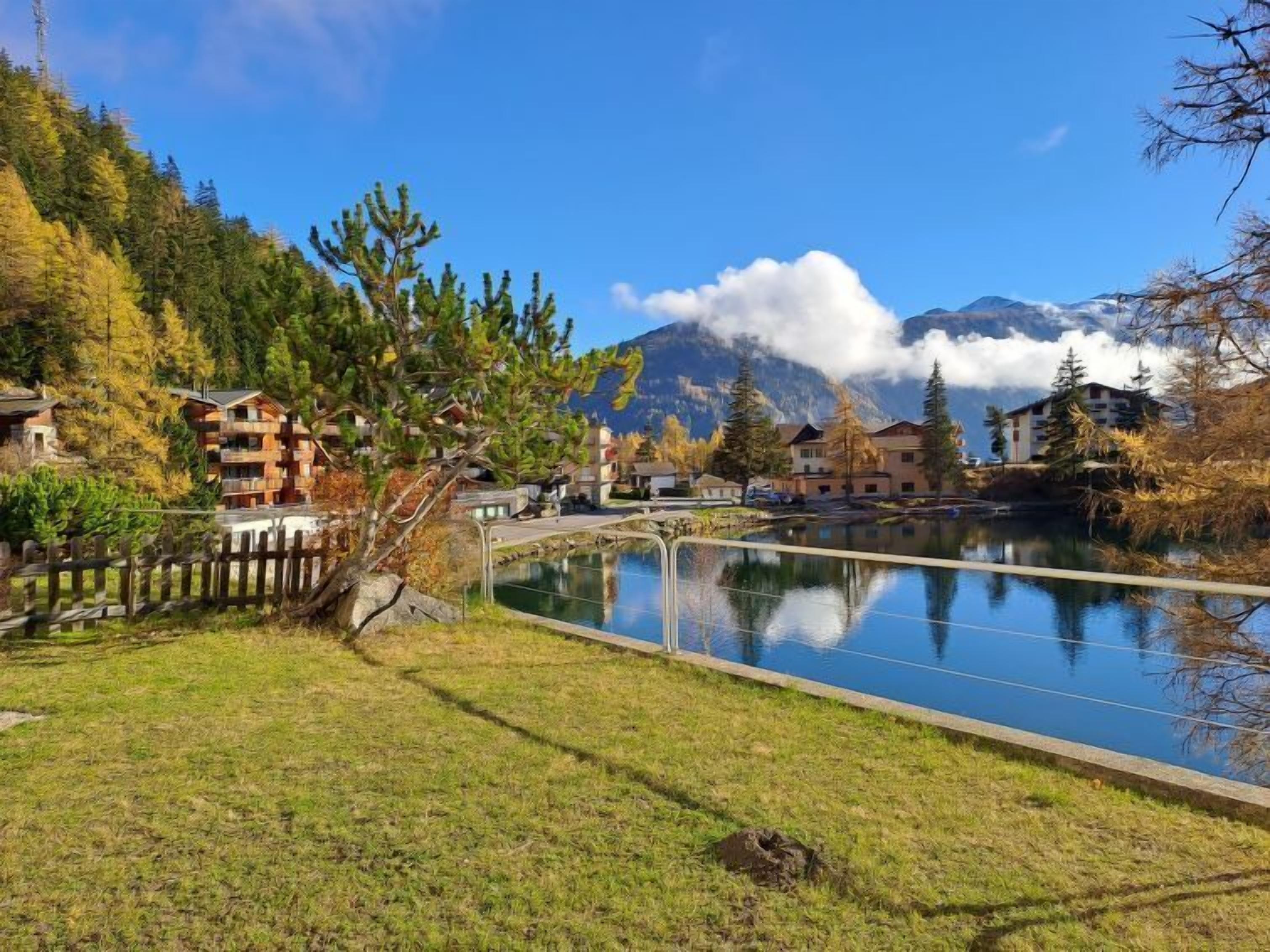 Vista a un lago con una cerca de madera, un estanque pequeño y edificios al fondo.