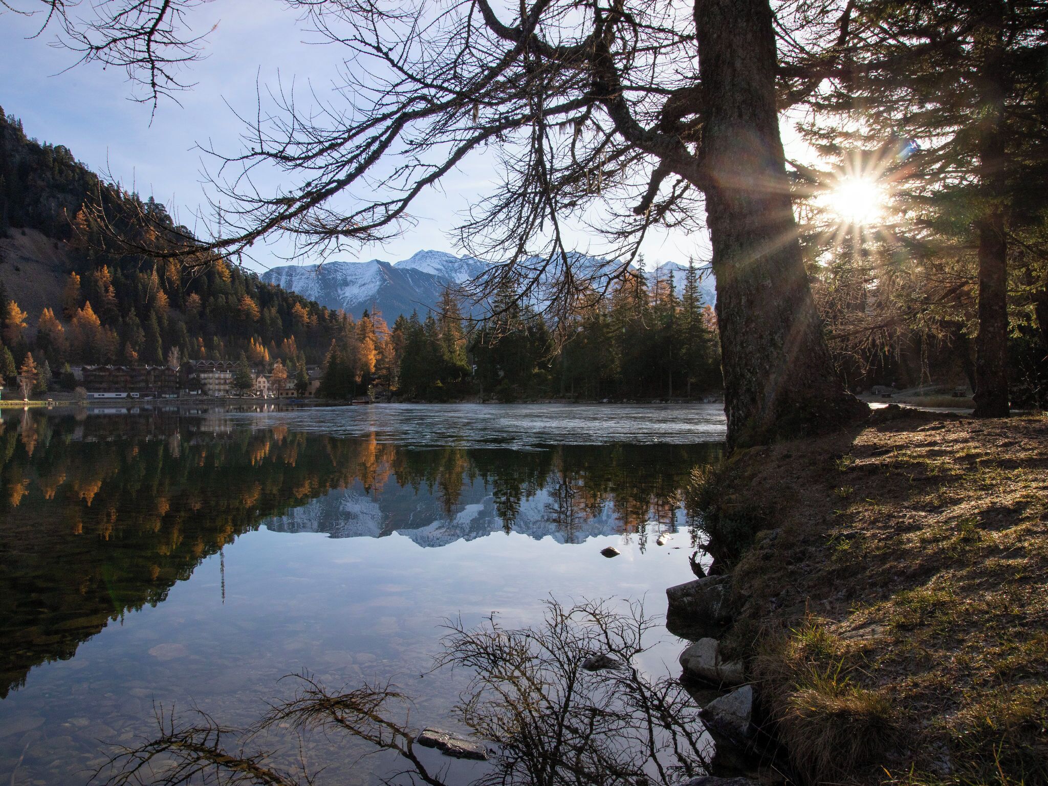 Ein ruhiger See mit Spiegelungen von Bäumen und Bergen, umgeben von einem bewaldeten Gebiet und einem Gebäudekomplex im Hintergrund.