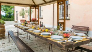 Une longue table en bois, dressée pour un repas sous une tonnelle, agrémentée de plantes en pot et de bouteilles de vin.