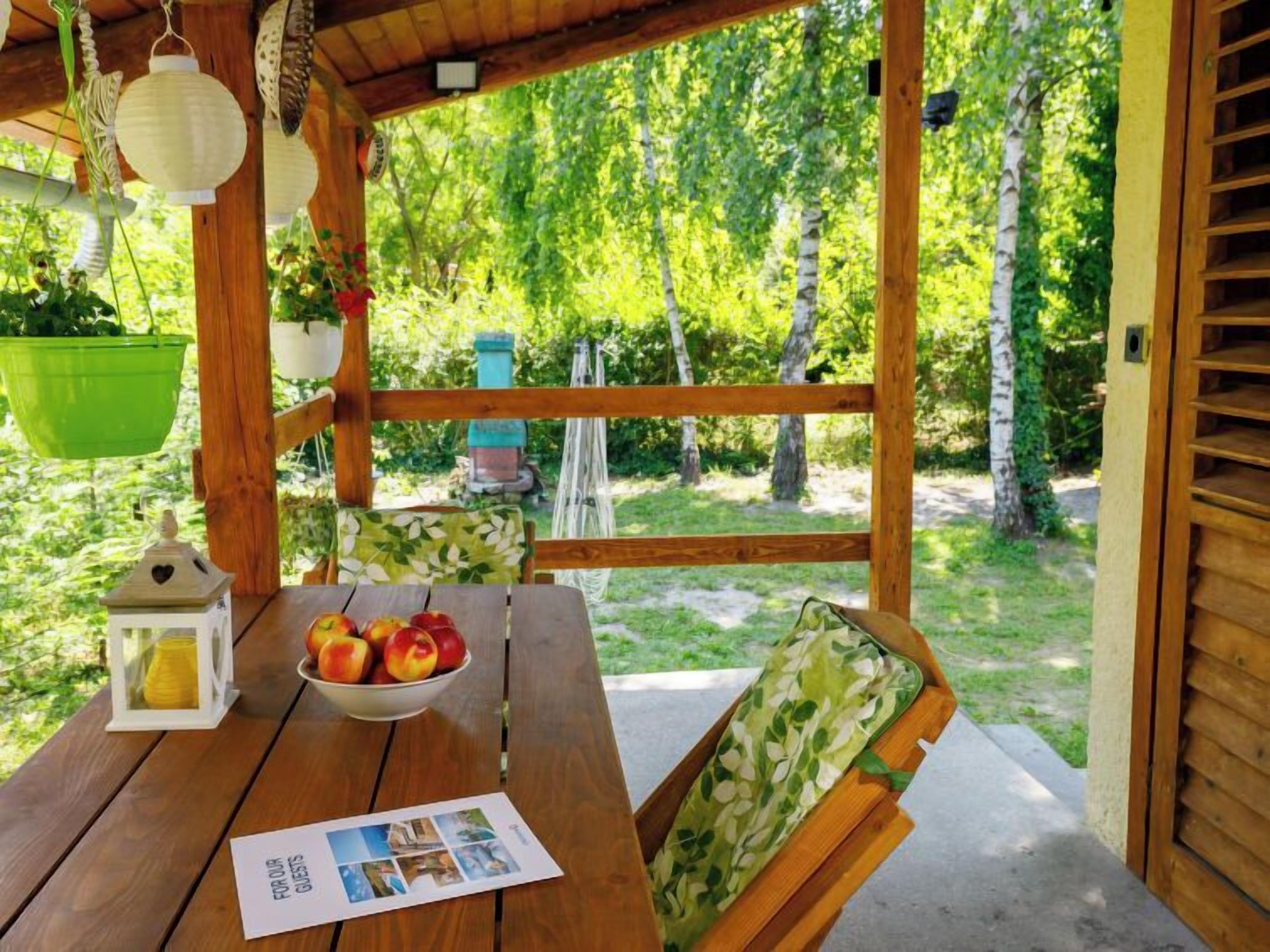 A wooden deck with a table, a bowl of apples, a lantern, and a chair with a leaf-patterned cushion.