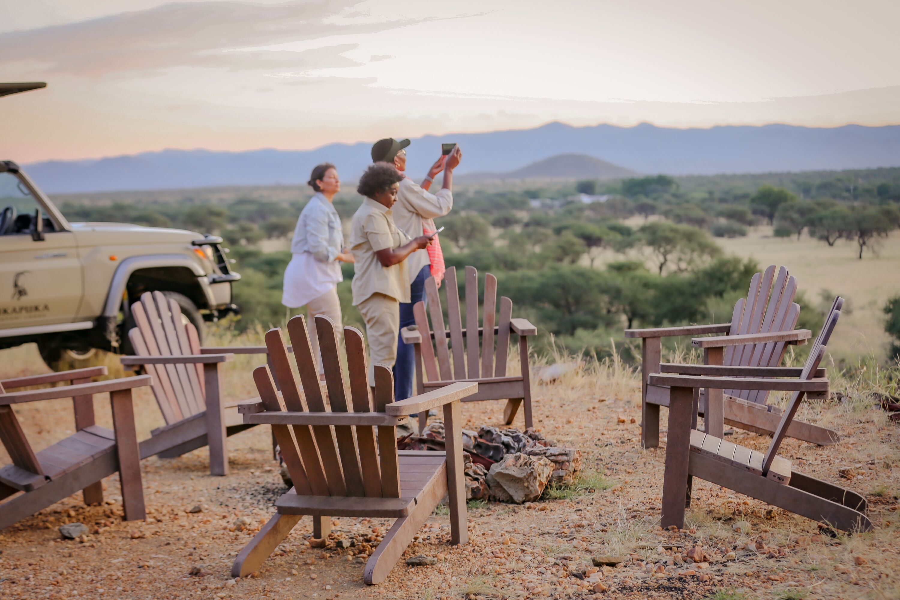 A group of people around wooden chairs and a vehicle in a natural setting with mountains in the background.
