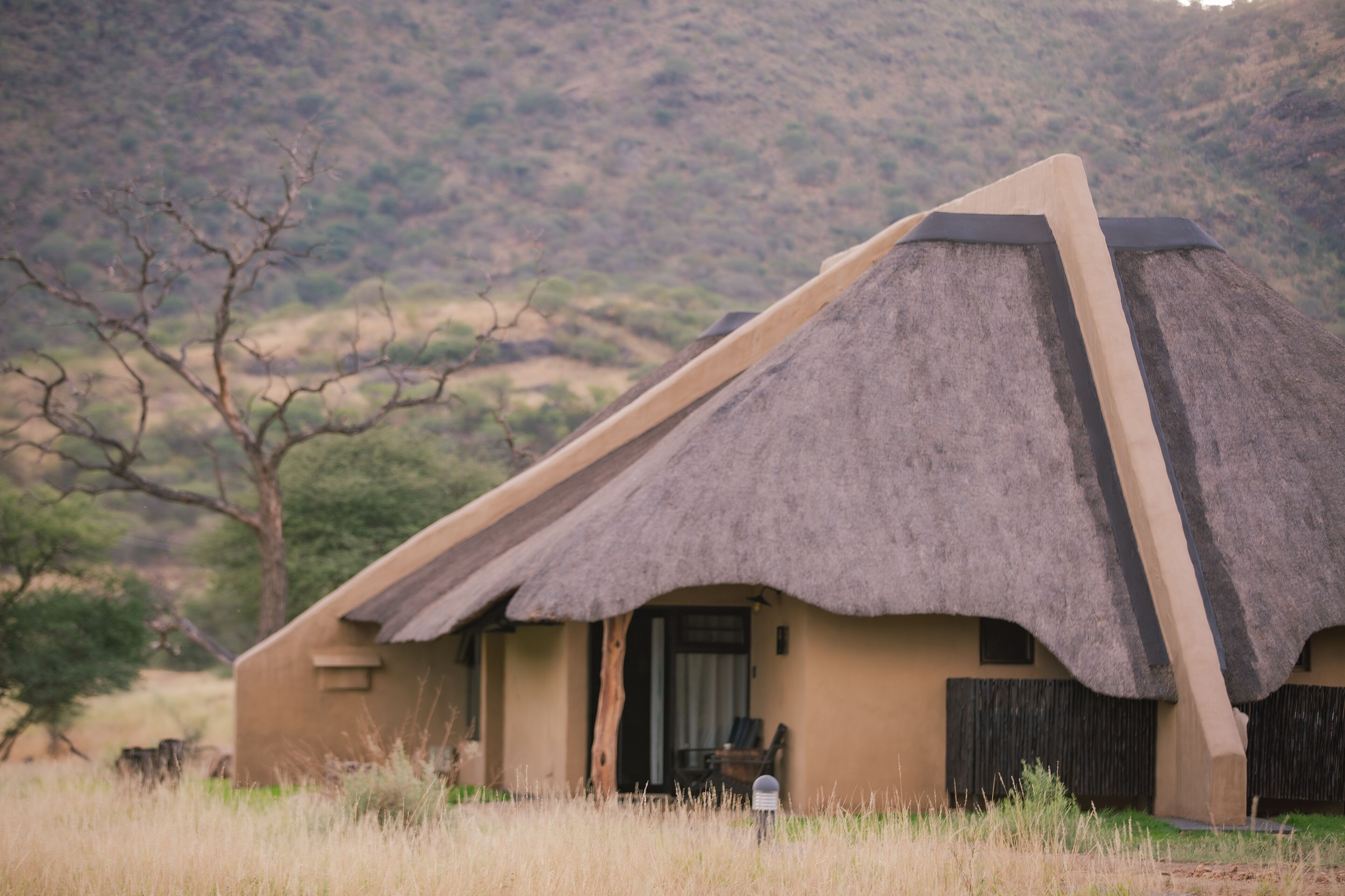 Un bâtiment à toit de chaume, doté d’une architecture simple et rustique, situé dans une zone herbeuse avec une colline en arrière-plan.