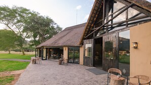 A building with a thatched roof, large glass doors, and a paved patio area with outdoor seating. - Okapuka Safari Lodge (Windhoek)
