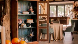 A dining area with a wooden cabinet displaying cakes and pastries, a round table with chairs, and a view of trees through a window. - Okapuka Safari Lodge (Windhoek)