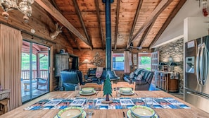 A rustic cabin interior with a wooden dining table set for four, a fireplace, leather seating, and a stone wall.