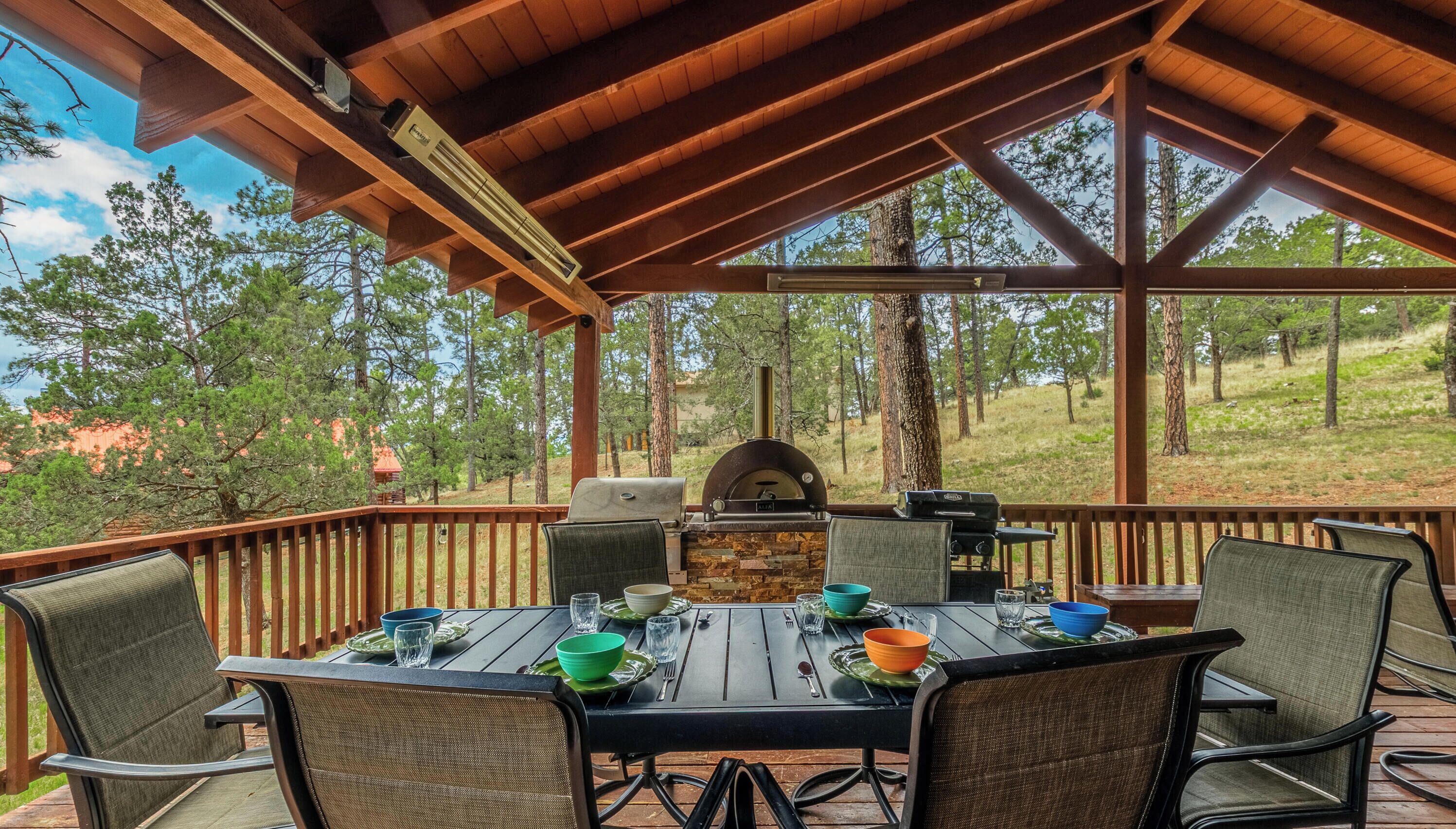 A covered outdoor dining area with a stone fireplace, wooden deck, and surrounding trees.