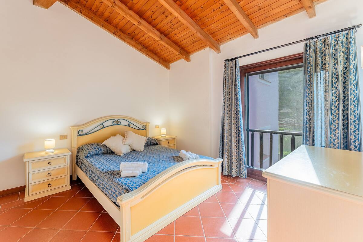 A bedroom with a bed, bedside tables, and a view through a window with curtains.