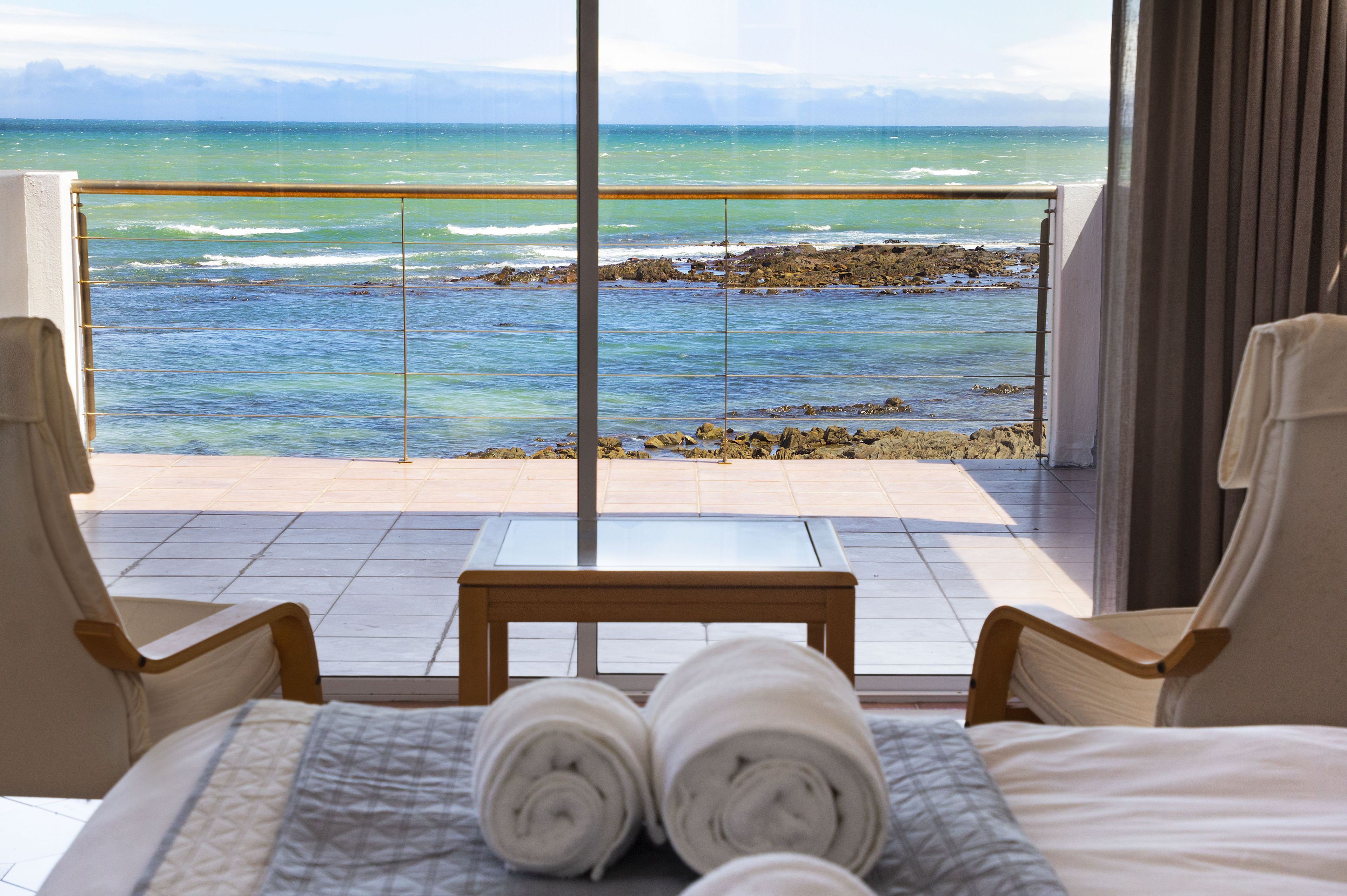 A hotel room with a bed, two chairs, a small table, and a view of the ocean through large windows.