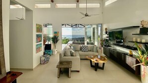 A spacious living room with a large sofa, a coffee table, and a TV. There are ceiling fans, a balcony with a view, and artwork on the walls.