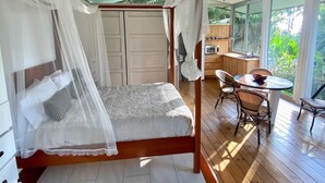 A bedroom with a canopy bed, a wooden floor, a dining area with a table and chairs, and a view of greenery outside.