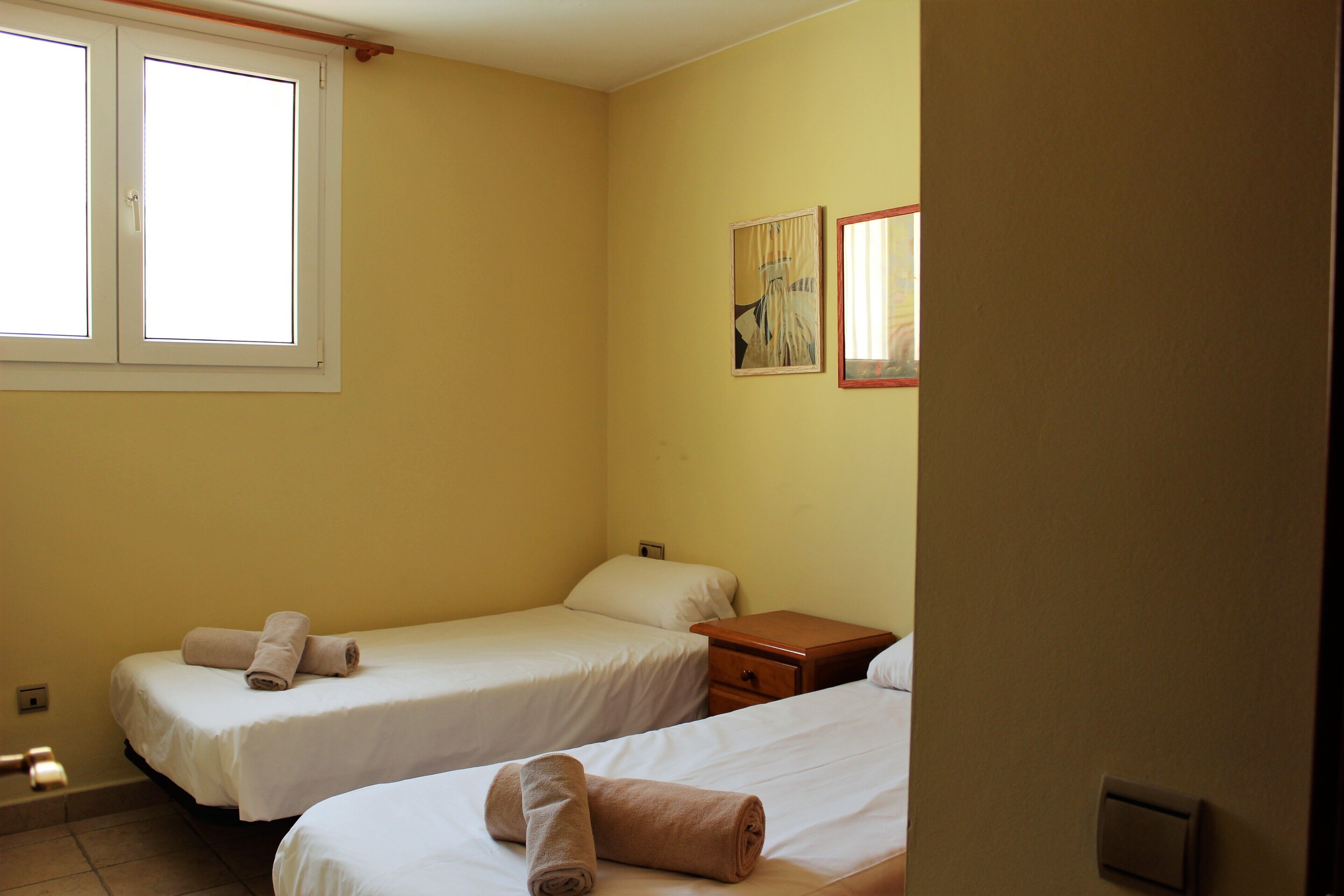 Ein Hotelzimmer mit zwei Einzelbetten, einem Holfnachtkästchen, einem kleinen Tisch und einem Fenster.
