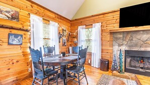 Interior acogedor de una cabaña con chimenea de piedra, paredes de madera, una mesa de comedor con sillas y un televisor montado en la pared.