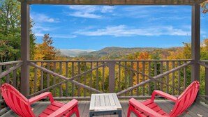 Uma área de estar coberta ao ar livre com duas cadeiras Adirondack vermelhas e uma mesa pequena, com vista para uma paisagem com árvores de cores outonais e colinas ao longe.