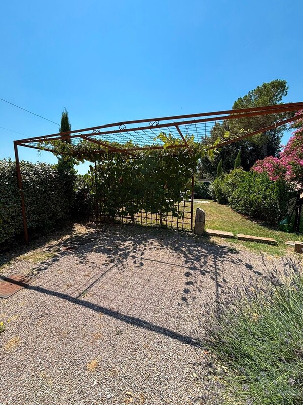 A gravel patio with a metal pergola structure, surrounded by greenery and a clear blue sky. - Vacation Home "Casa di Campagna" with Private Terrace & Wi-Fi (Colle Val d'Elsa)
