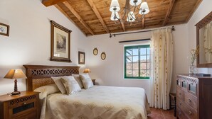 Ein Schlafzimmer mit Holz-Kopfteil, einem Bett mit gemusterten Textilien, einem Nachttisch mit Lampe, einer Kommode, einem Fenster mit Vorhängen und einer Chandelieber.