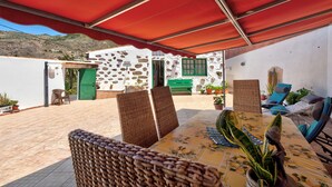 Eine Terrasse mit Korbstühlen, einem Tisch mit Blumenmuster-Tischdecke und einem roten Sonnensegel.