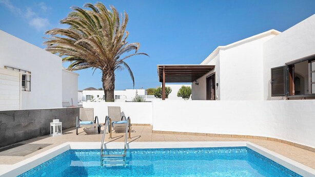 Une piscine avec un palmier, un espace salon abrité et un bâtiment blanc.
