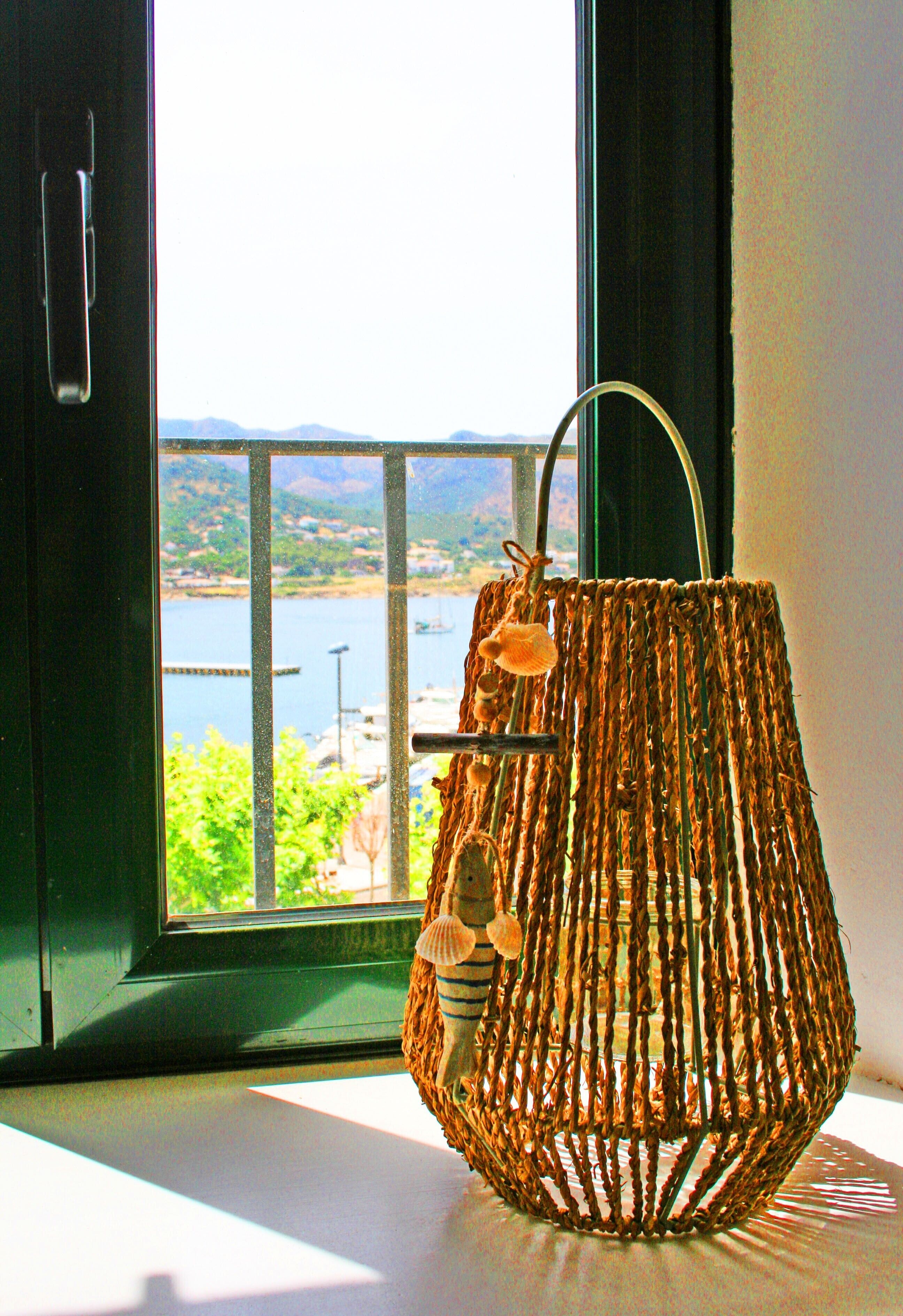 Un panier tressé, doté de poignées, est posé sur un rebord de fenêtre, d’où l’on aperçoit une étendue d’eau et des terres lointaines.