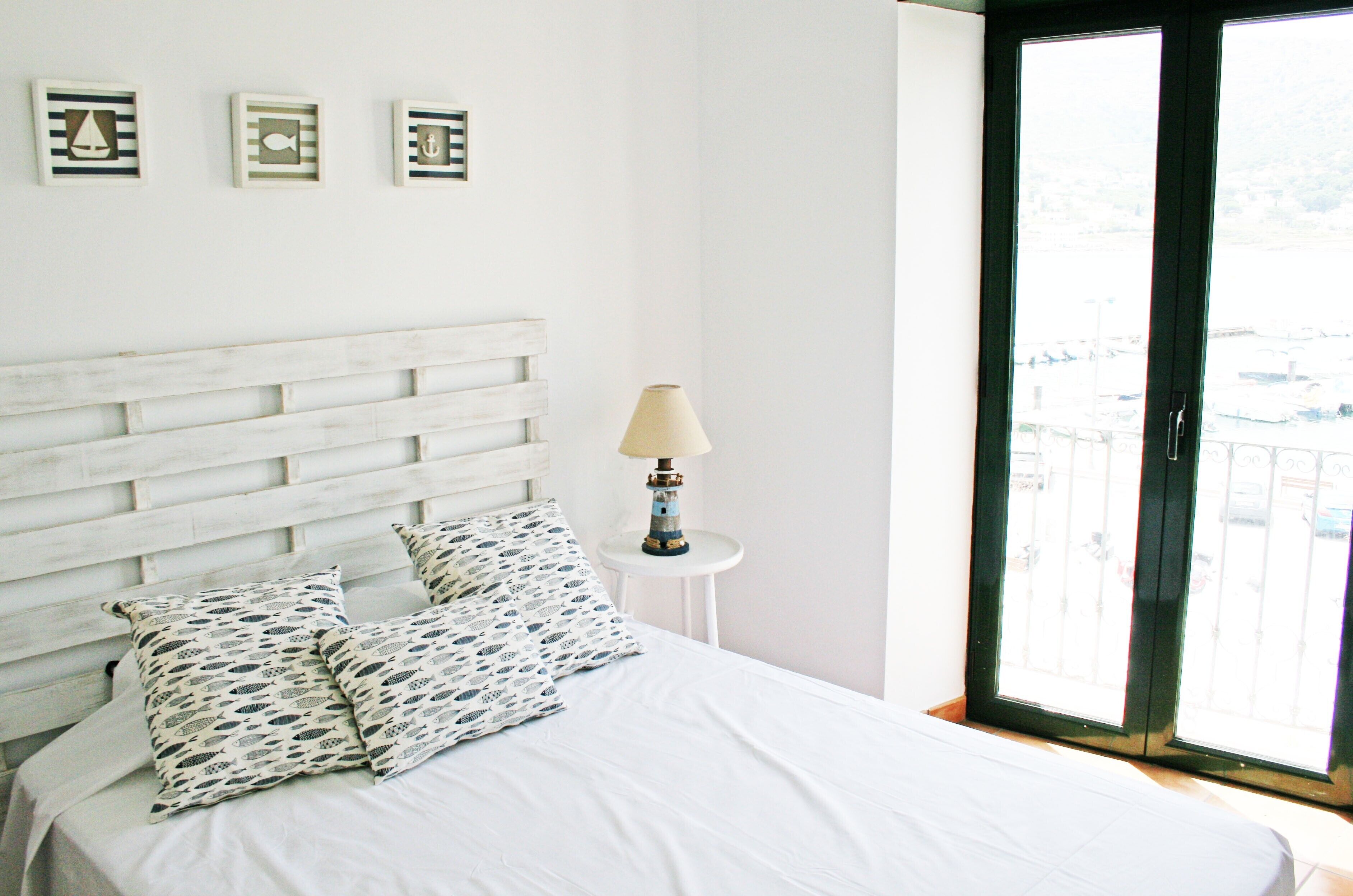 Een bed met witte lakens en gestreepte kussens, een bedlamp en een raam met uitzicht op een balkon.