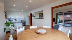 A modern kitchen with white cabinets, a dining table with chairs, and a large window with a view of a brick building. - Montaview - Mumbulla St 24 (Bermagui)