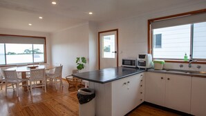 A kitchen with white cabinets, a black countertop, a microwave, and a sink. There is a dining table with chairs and a potted plant. - Montaview - Mumbulla St 24 (Bermagui)