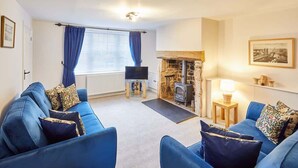 A living room with blue sofas, a stone fireplace, a television, and a window with curtains. - Host & Stay | Kenmore (Hexham)