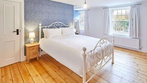 A bedroom with a white bed, wooden flooring, a blue patterned wall, a window with curtains, and a nightstand with a lamp. - Host & Stay | Kenmore (Hexham)