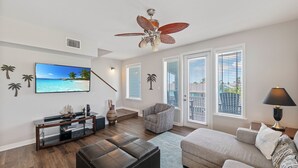 A living room with a TV, sofa, ottoman, ceiling fan, and a view through the window. - Three story townhome. Garage access. Golf cart included. Very short drive to the (Port Aransas)