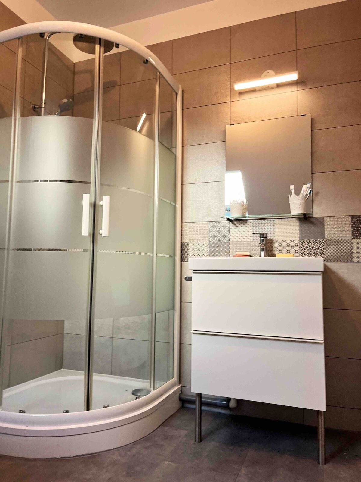 Et moderne badeværelse med et glasafskærmet brusebad, et hvidt badeværelsesmøbel med skuffer og et flisebeklædt bagpanel.