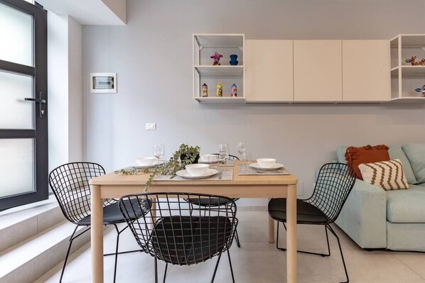 Un comedor con una mesa dispuesta para cuatro personas, rodeada de sillas negras con estructura metálica. Encima de la mesa, un armario blanco con estantes, y en el fondo, un sofá azul claro con un cojín estampado.