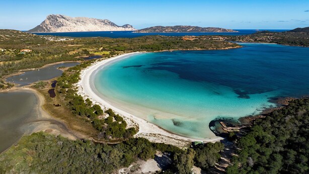 Vue aérienne d’une zone côtière avec une plage, des eaux turquoise et limpides, et un paysage montagneux en arrière-plan.