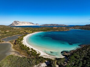 Ovenudblik over et kystområde med en strand, klart turkis vand og et bjergrigt bagland.