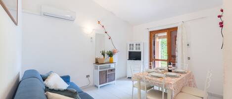 Una sala de estar con un sofá azul, una mesa de comedor preparada para cuatro personas y una cocina con televisor y armarios.