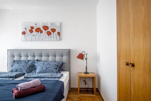 A bedroom with a tufted headboard, a bed with blue bedding, a red towel, a small wooden nightstand with a lamp, and a wooden wardrobe.