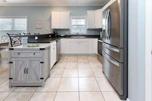 A kitchen with white cabinets, a stainless steel refrigerator, and a window with blinds. - 2nd Row 5 BR Beach House with Private Pool - Hip Nautic (Surfside Beach)