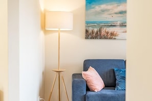 A room with a standing floor lamp, a blue sofa with two pillows, and a beach-themed painting on the wall.