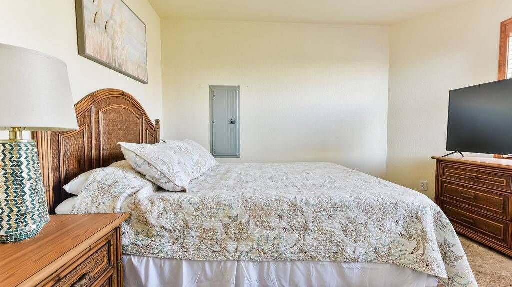 Une chambre à coucher avec une tête de lit en bois, un lit recouvert d’une couvre-lit à motifs, une table de chevet avec une lampe, une commode et un téléviseur posé sur un meuble en bois.
