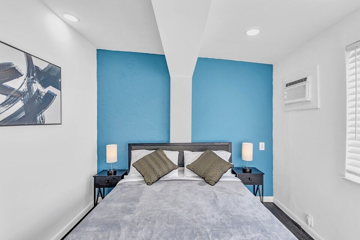 A modern bedroom with a bed, two nightstands, a wall-mounted air conditioning unit, and a window with blinds.