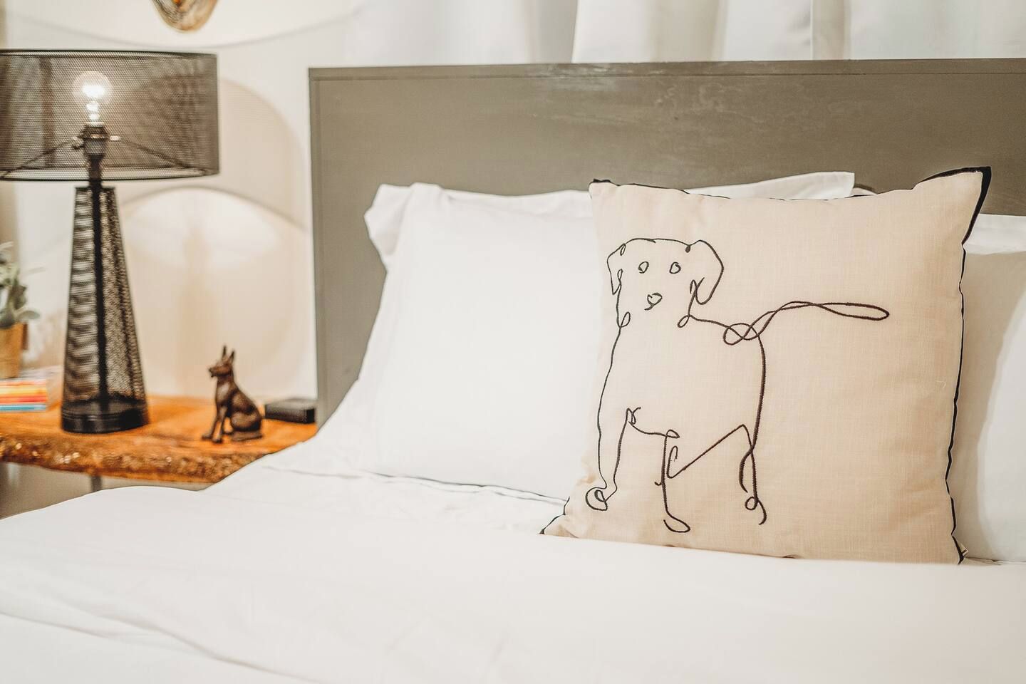 A bed with white bedding, a beige pillow with a line drawing of a dog, a lamp, and a small figurine on a wooden side table.
