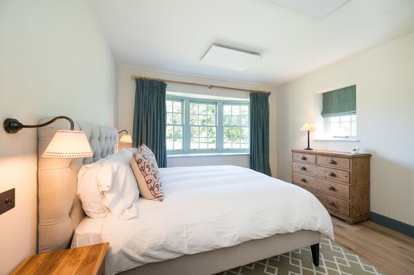 A bedroom with a large bed, a dresser, a nightstand, a lamp, and a window with curtains.