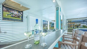 Eine Bar mit einem Fernseher, der ein Sportspiel zeigt, einer Flasche The Beachhouse Chardonnay Blanc und zwei Gläsern Weißwein.