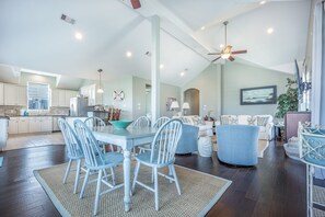 Un vaste espace de vie comprenant une table à manger, des chaises et un canapé. Au fond, on aperçoit une cuisine avec des meubles blancs et une fenêtre.