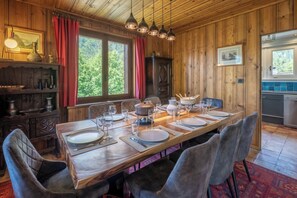 Een houten eetkamer met een lange tafel gedekt voor zes personen, omringd door stoelen, en uitzicht op de buitenwereld door grote ramen.