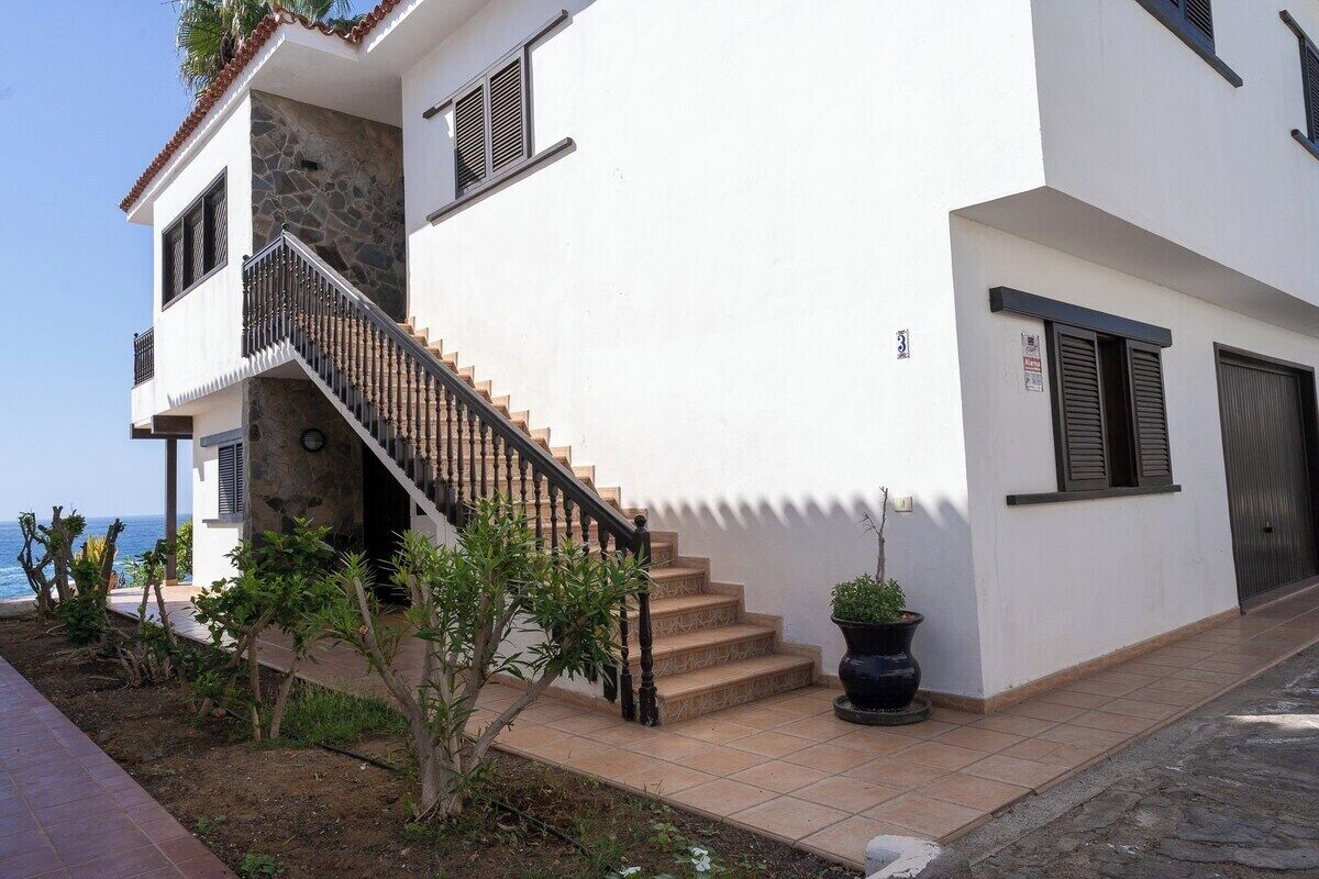 A two-story building with a staircase leading to the upper floor, a balcony with a view of the sea, and a potted plant on the ground floor.