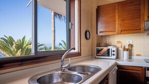 A kitchen with a double sink, wooden cabinets, a window with a view of palm trees, a toaster, and a clock on the wall. - Ocean front 3 bedroom apartment (Santiago del Teide)