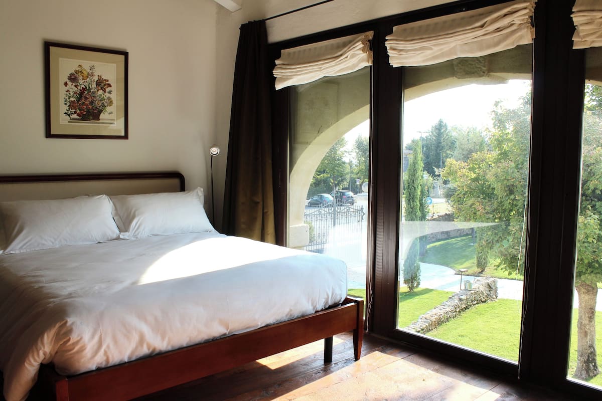 A bedroom with a large bed, a framed floral artwork on the wall, and a view of a garden through a glass door.