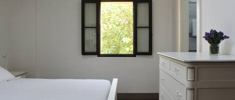 Un dormitorio con una cama, un tocador, una ventana con contraventanas y un florero con flores.