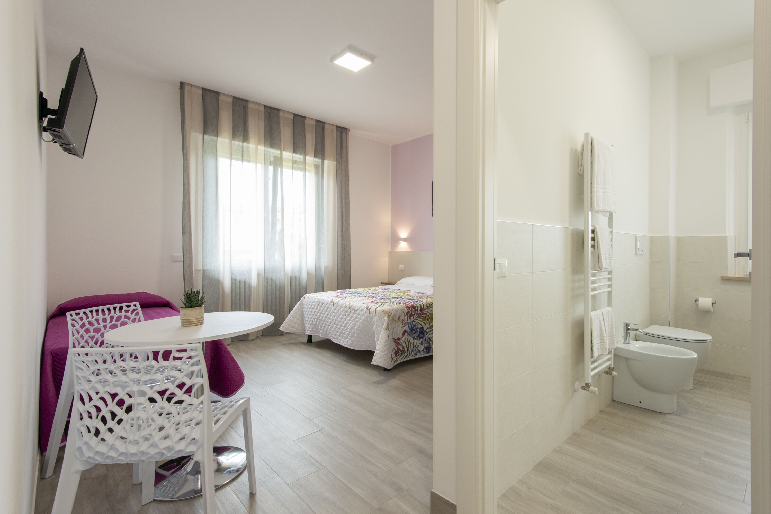 Et moderne hotelværelse med seng, et lille bord med stole, et tv ophængt på væggen og et badeværelse med toilet og vask.